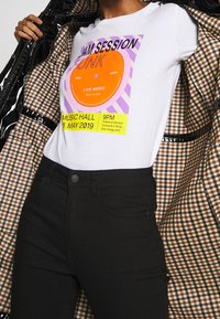 Person trägt schwarze Hose und ein weißes T-Shirt mit Aufdruck eines Schallplattendesigns sowie Text zu einem Musikevent und hält einen geöffneten karierten Mantel.