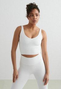 Biely rebrovaný crop top so širokými ramienkami a hlbokým výstrihom do V. V páre s vysokými legínami, ktoré sú v rovnakom štýle, obidva s hladkou textúrou.