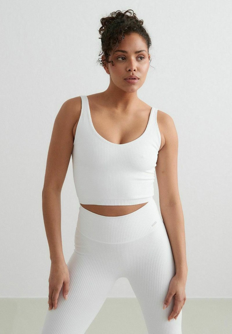 Biely rebrovaný crop top so širokými ramienkami a hlbokým výstrihom do V. V páre s vysokými legínami, ktoré sú v rovnakom štýle, obidva s hladkou textúrou.