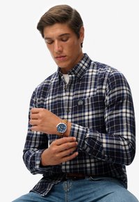 Marineblauw-wit geruit overhemd met knopen en een borstzakje, gecombineerd met een blauw horloge en lichtblauwe spijkerbroek, gedragen door een model dat het horloge verstelt.