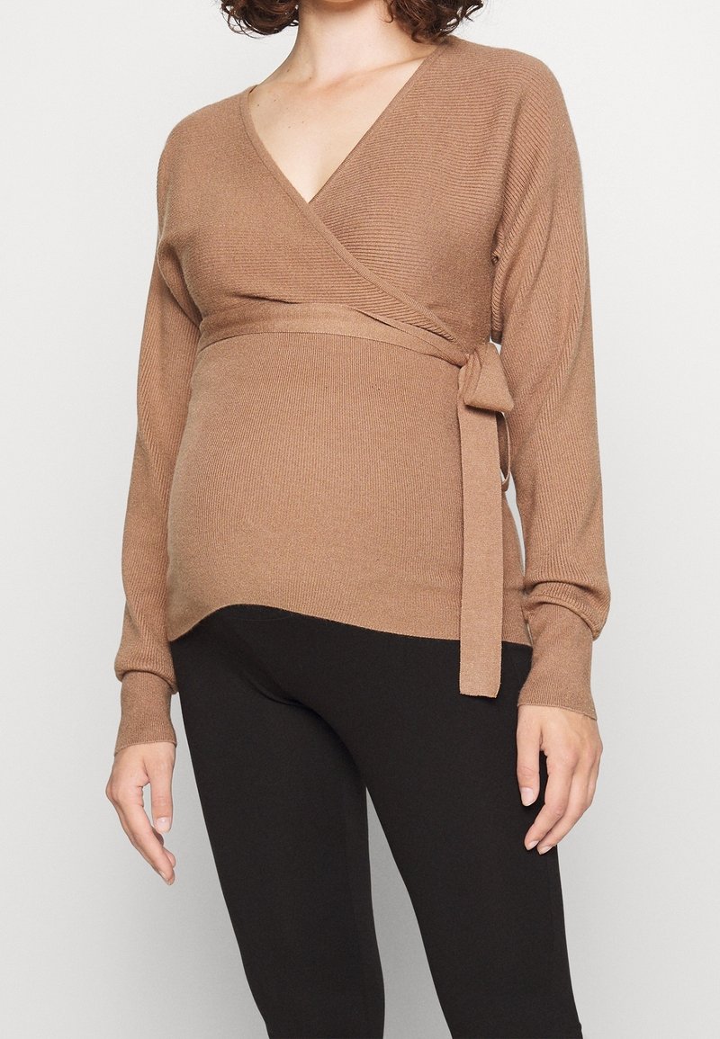 Femme portant un pull beige croisé noué sur le côté et des leggings noirs, debout devant un fond clair uni.