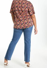 Persona che indossa jeans blu e una camicia a maniche corte con stampa floreale, in piedi con una gamba leggermente piegata, indossando scarpe con tacco alto bianche.