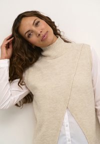 Pull en tricot sans manches beige à col roulé, avec une finition texturée et un design enveloppant, superposé sur une chemise blanche à col.