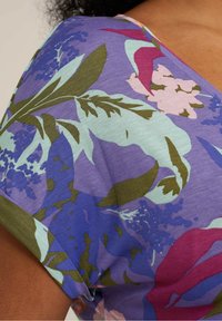 Květinový top v purpurové barvě s zelenými a růžovými květinami. Tkanina vypadá měkce s hladkým povrchem a krátkými rukávy.