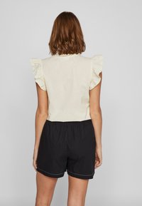 Top bege claro com mangas em babados, combinado com calções pretos com uma cintura elástica e uma bainha branca cosida.