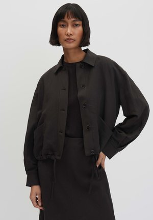 Femme aux cheveux courts noirs porte une veste noire boutonnée sur un haut côtelé noir avec une jupe assortie, posant devant un fond uni.