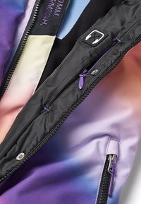 Gros plan sur une veste dégradée multicolore avec des fermetures éclair violettes, une doublure intérieure noire, un curseur de fermeture éclair et un patch en forme d'icône d'écouteurs blanc.