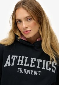 Mujer rubia con ojos verdes que lleva una sudadera negra con el texto "ATHLETICS SD. UNIV. DPT" y una camiseta de rayas rosas debajo.