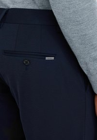 Donkerblauwe broek van gladde stof, met een zijzak met knoopsluiting en een klein metalen merkteken op de achterkant.