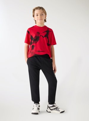 Ragazzo in piedi con le mani nelle tasche, indossa una maglietta da calcio rossa con grafiche, pantaloni della tuta neri e scarpe da ginnastica bianche su sfondo bianco.