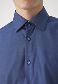 Modrá košile na knoflíky s texturovanou tkaninou, opatřená klopovým límečkem a odpovídajícími knoflíky. Přiléhavý střih a jemný lesk.