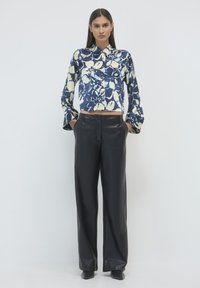 Femme debout portant une chemise boutonnée florale bleu marine et crème et un pantalon en cuir noir à taille haute et jambes larges, les mains dans les poches.