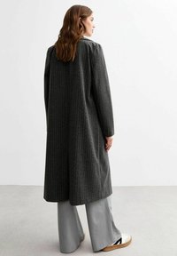 Abrigo largo de color gris oscuro con rayas verticales, corte ajustado, diseño de un solo botón, combinado con pantalones de pierna ancha de color gris claro y calzado elegante.