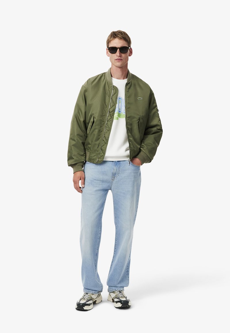 Giubbotto bomber verde oliva realizzato in materiale liscio, con chiusura a zip frontale e polsini a coste, abbinato a jeans denim azzurri e scarpe da ginnastica con motivo.