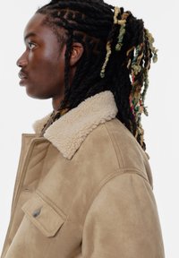 Tan ruskindsjakke med en cremet faux fur-krave, sidelomme og knappedetalje. Model vist i profil med langt, tekstureret hår.
