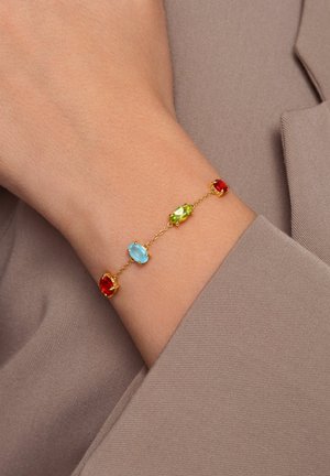 Goldarmband mit roten, grünen und blauen ovalen Edelsteinen, die in Krappen gefasst sind und durch eine feine Kette verbunden werden, getragen an einem nackten Handgelenk über einem Stoff.