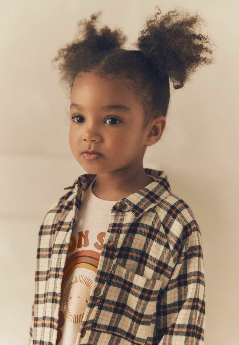 Jaunas vaikas su garbanotais plaukais, surištais į dvi pūkuotas šukuosenas, vilkintis languotą marškinį virš kūno spalvos marškinėlių su piešiniu, žvelgiantis šiek tiek į šoną prieš neutralią foną.