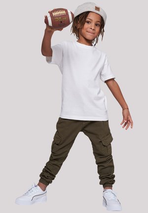 Child in a white t-shirt, olive green cargo pants with side pockets, gray knit hat, holding a brown football, wearing white sneakers.