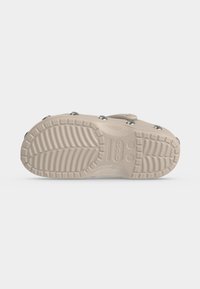 Σόλα ενός μπεζ Crocs slip-on clog που δείχνει το σχέδιο πέλματος, τις σημάνσεις μεγέθους και τις στρογγυλές μεταλλικές τρουκς στις άνω πλευρές.