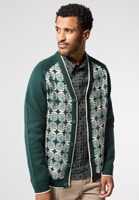 Zelený cardigan s vzorovaným předním dílem s kružnicemi v bílé a tyrkysové barvě, žebrovanými manžetami a knoflíky ladícími s barvou těla.