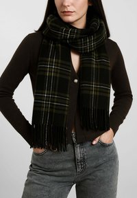 Sciarpa a quadri neri e verdi con bordi sfrangiati, realizzata in materiale morbido. Indossata in modo sciolto intorno al collo, abbinata a un cardigan scuro.