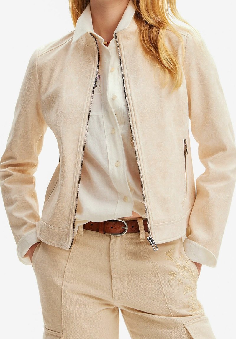 Lichtbeige suède jas met rits, minimale kraag en zijzakken. Gepaard met een wit shirt en lichtbeige broek met bloemendecoratie.