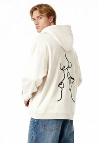 Sudadera de algodón en color crema con capucha ajustable. Presenta un diseño de arte lineal negro en la parte posterior. Ajuste relajado y puños acanalados.