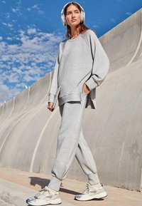 Grijze oversized sweatshirt en bijpassende sweatpants gemaakt van zachte stof. Model draagt zilveren sneakers en een hoofdtelefoon, staande buiten.