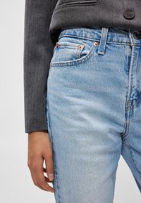 Világoskék farmernadrág enyhe mosott hatással, vörösréz szegecsekkel és egyenes szabású kialakítással, sötétszürke blézerrel viselve.