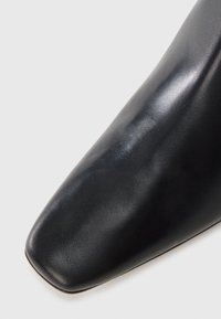 Botim de tornozelo em pele preta com biqueira apontada, textura lisa e design elegante. A parte superior tem um acabamento brilhante sem adornos visíveis.