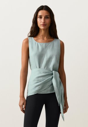 Žena s dlhými tmavými vlasmi, ktorá má na sebe svetlomodrý tielkový top s viazaným detailom v páse a čierne nohavice, stojaca na jednoduchom pozadí.