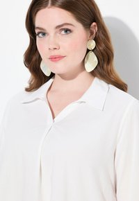 Camicia bianca con colletto, leggermente lucida, dotata di una fila di bottoni. Orecchini ovali giallastri traslucidi di grandi dimensioni con una finitura naturale.