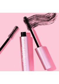 Roze mascarabuis met het label "Better Than Sex" en een zwarte borstel, naast een gebogen streek van zwarte mascara op een roze achtergrond.