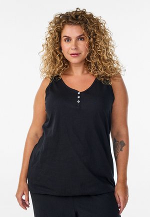 Kvinde med krøllet blondt hår iført en sort ærmeløs top med tre knapper og sorte bukser, der viser en rosetatovering på venstre underarm.