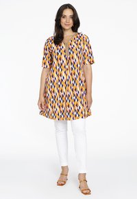 Tunique colorée avec des motifs de diamants orange, violet et jaune ; manches courtes ; décolleté en V ; associée à un pantalon blanc et des sandales marron.