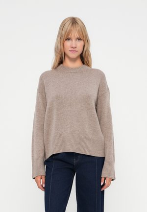 Beige strikkegenser med rund hals, ribbede mansjetter og hem. Løs passform med cropped lengde, kombinert med mørke denim jeans.