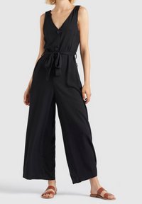 Black sleeveless jumpsuit with a V-neck, buttons, and a tied belt at the waist. Flared legs and soft fabric texture. Worn with tan sandals.