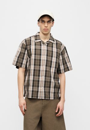 Jeune homme portant une casquette beige LA, une chemise à manches courtes boutonnée à carreaux marron et blanc, et un pantalon marron ample, debout devant un fond blanc.