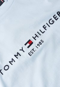Light blue cotton T-shirt featuring embroidered black text: "TOMMY HILFIGER" and "EST. 1985," with a red, white, and blue logo accent.