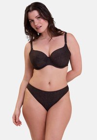 Femme aux cheveux foncés portant un soutien-gorge pailleté brun foncé assorti et des culottes taille haute sur un fond blanc uni.