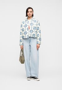 Γυναίκα που φοράει μπλε και λευκό μπουφάν με μοτίβο, ανοιχτόχρωμο τζιν με φαρδιά πόδια, μαύρα αθλητικά παπούτσια και κρατά μια μικρή ελιά τσάντα χειρός.