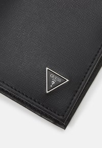 Portefeuille noir texturé avec une quincaillerie argentée triangulaire présentant le logo "GUESS", des bords cousus et une finition lisse. Design compact.