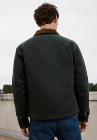 Dunkelgrüne Jacke mit einem Cordkragen, die eine glatte Textur aufweist, Knöpfe an den Manschetten hat und einen leicht abgerundeten Saum besitzt. Getragen mit blauen Jeans.