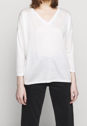 Vrouw draagt een losse witte V-hals met lange mouwen en zwarte broek, staand tegen een effen lichtgrijze achtergrond.