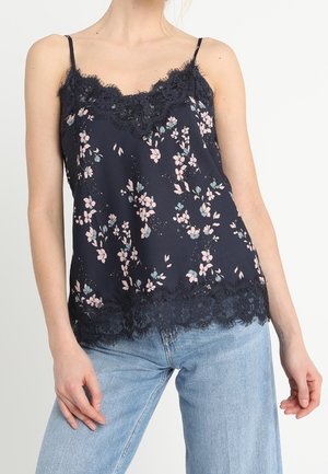 Kobieta nosząca granatowy kwiatowy top z koronkową lamówką, w połączeniu z jasnoniebieskimi jeansami, stojąca na jednolitym tle.