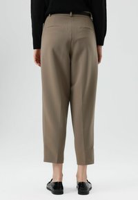Pantalon beige sur mesure avec une longueur raccourcie à la cheville, doté de deux poches arrière et d'une texture lisse. Associé à des chaussures noires sans lacets.