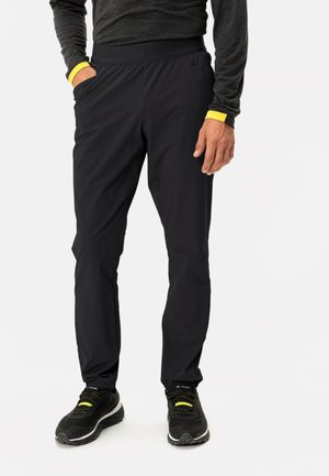 Zwarte broeken met een taps toelopend ontwerp, elastische tailleband, zakken en gele accenten op de manchetten. Draagt zwarte sportschoenen.