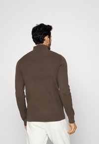 Pull marron côtelé avec un col montant et des manches longues, présentant un design ajusté et un tricot texturé. Détails de couture visibles au niveau de l'ourlet.