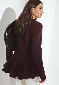 Bordový pletený sveter s rozšíreným lemom, dlhými rukávmi a textúrovanou látkou. Čierna okrúhla kabelka spočíva na ruke.