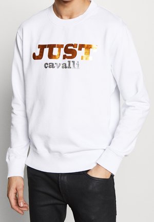 Mand iført en hvid sweatshirt med metallisk bronze "JUST" og sølvfarvet "cavalli" trykt på brystet, kombineret med sorte bukser.